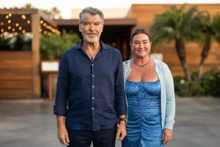 Pierce Brosnan (in a navy blue button-down and necklace) and wife Keely Shaye Smith (in a blue patterned dress and light cardigan) smiling together outside a restaurant.