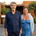 Pierce Brosnan (in a navy blue button-down and necklace) and wife Keely Shaye Smith (in a blue patterned dress and light cardigan) smiling together outside a restaurant.