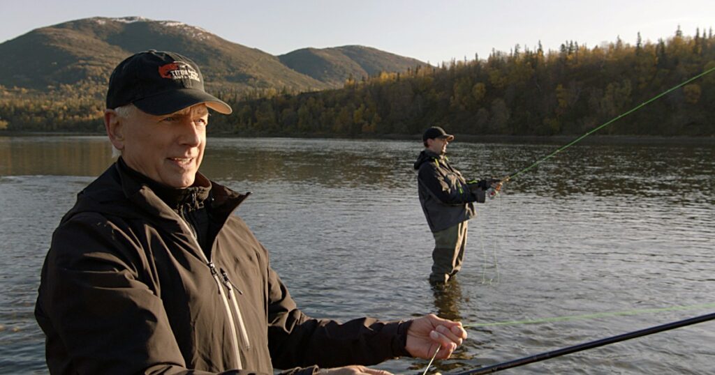 Mark Harmon as Leroy Jethro Gibbs in a black cap and jacket, smiling as he fly-fishes on an Alaskan river in NCIS